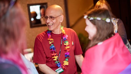 A smiling man wearing a rainbow-coloured flower garland around his neck is talking to people, Birmingham Back-to-Backs